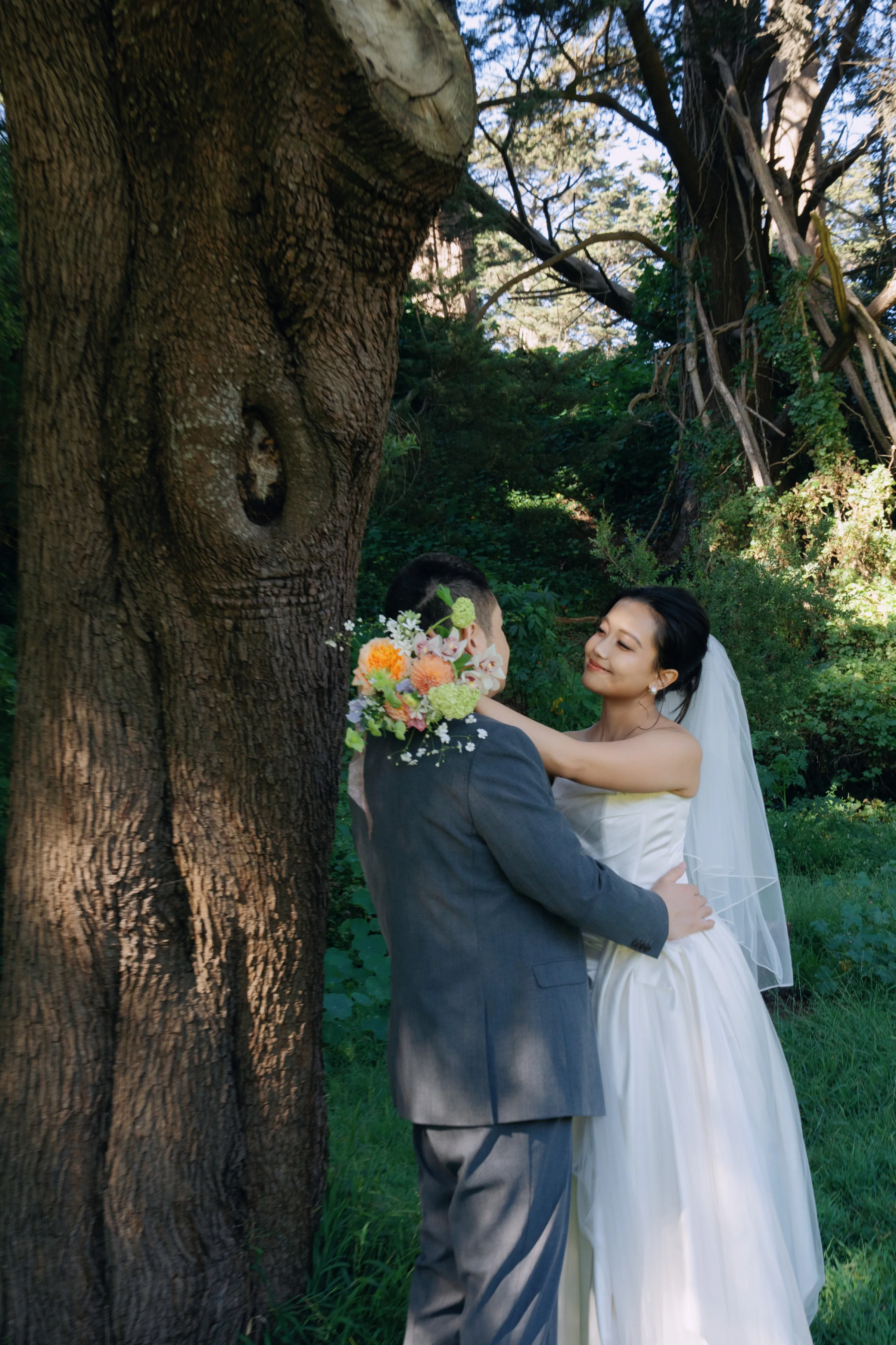 Couple embracing beside a large tree trunk, the bride holding a bouquet