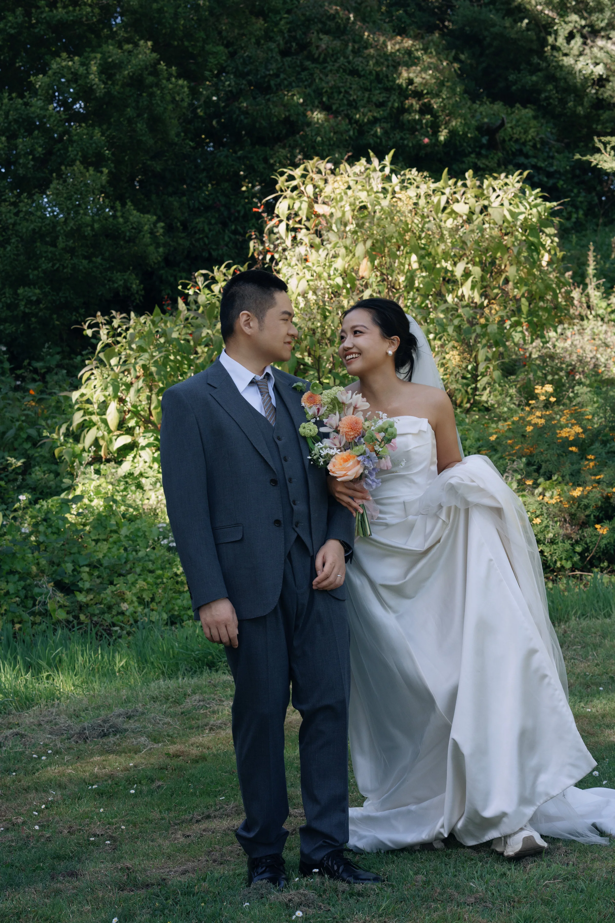 Couple standing close among garden greenery, the bride holding a peach bouquet
