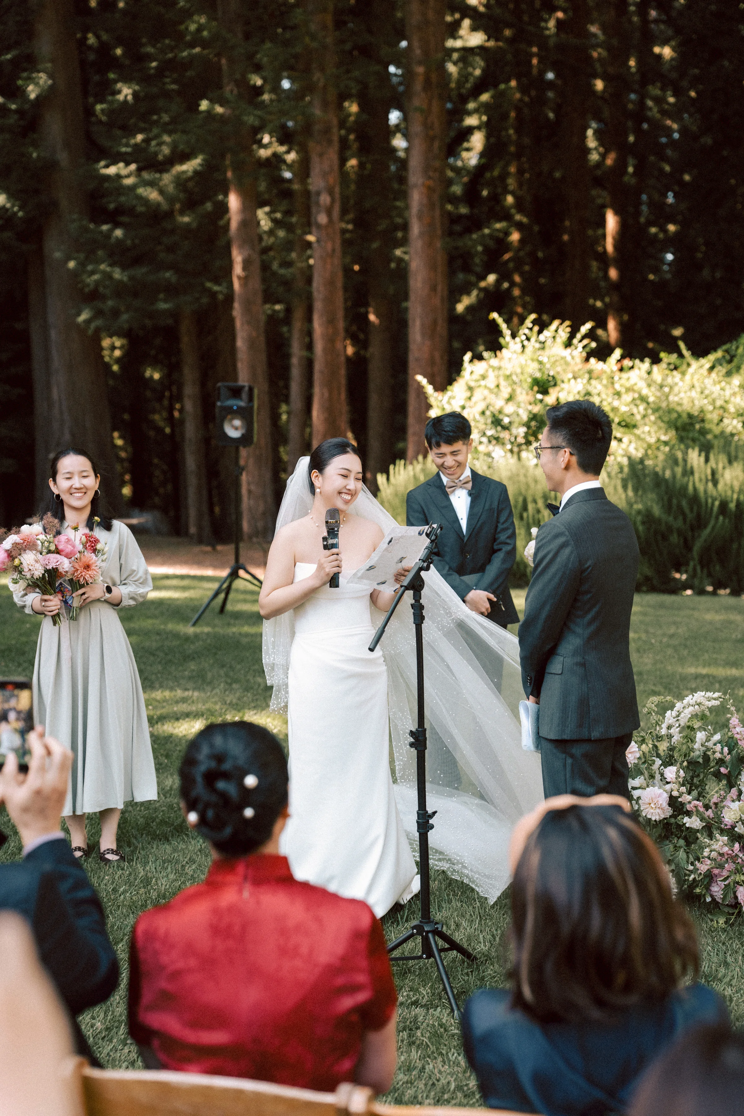 Couple exchanging vows at a microphone in the redwood grove as a guest films