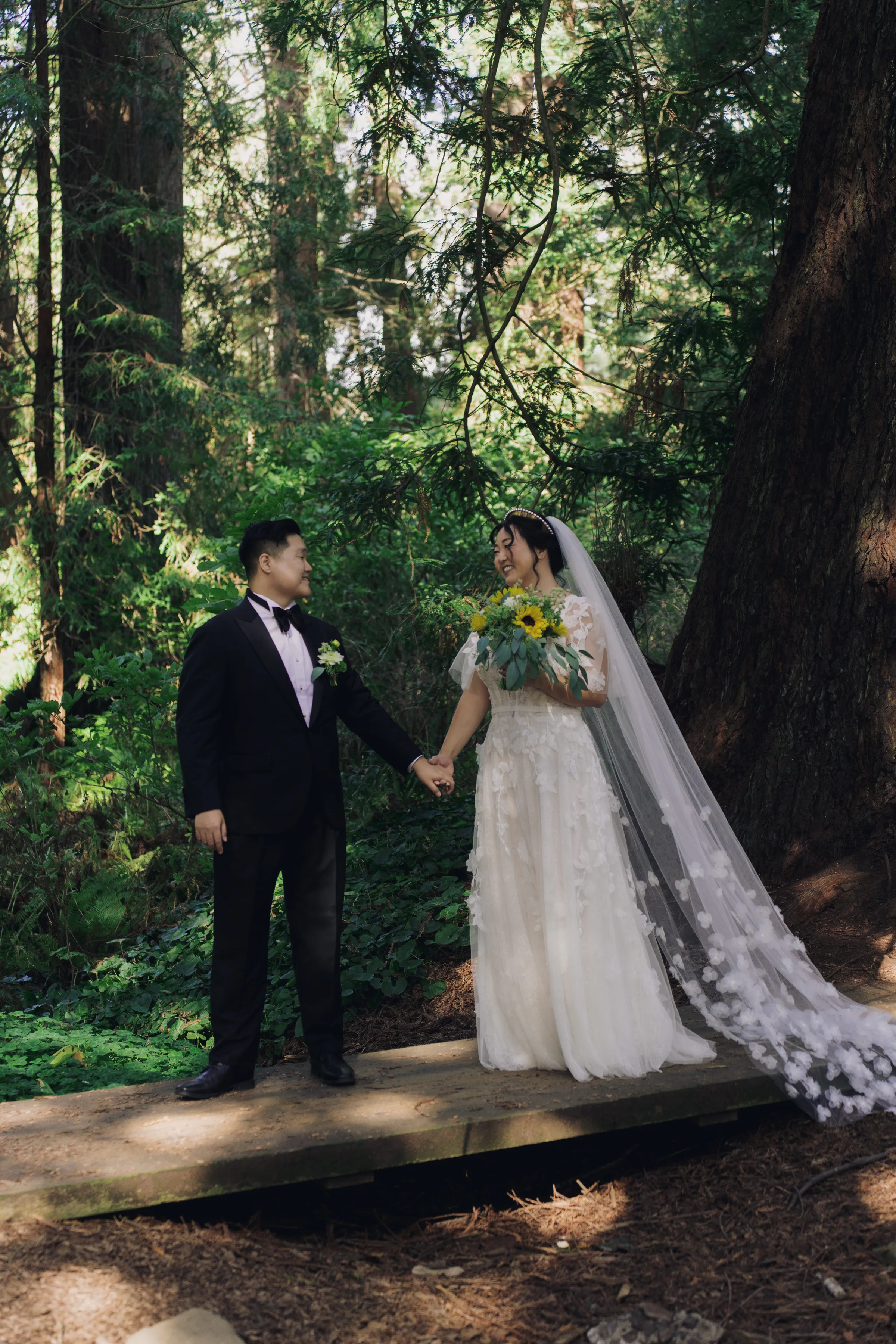 Newlyweds hold hands beneath towering trees, with the bride carrying a sunflower and greenery bouquet.