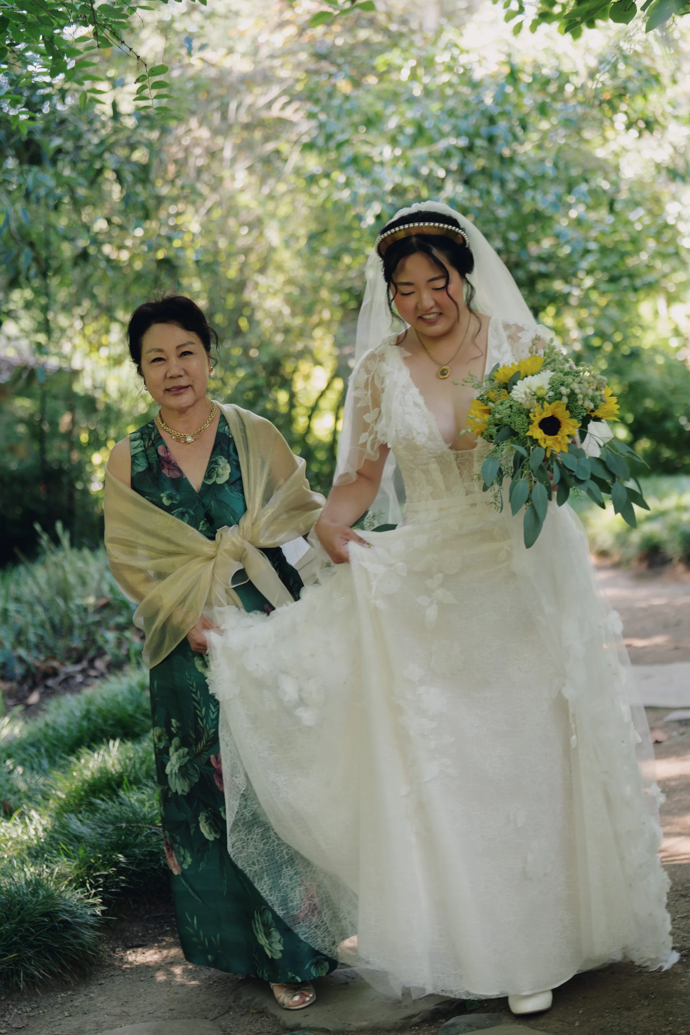 Bride walks along a garden path with a woman in a green floral dress while holding her sunflower bouquet.