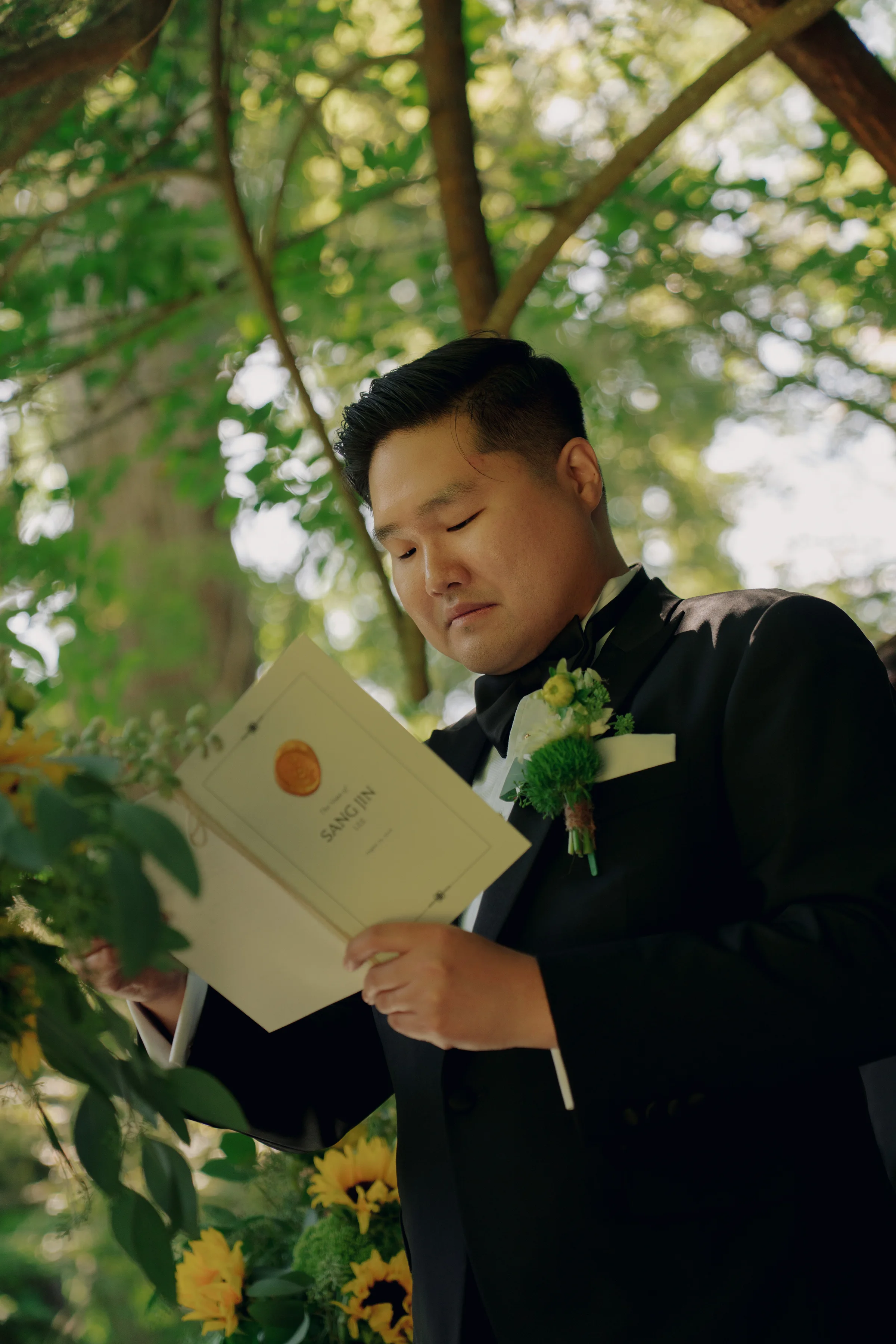 Groom reads a letter beside sunflower ceremony arrangements beneath leafy garden branches.