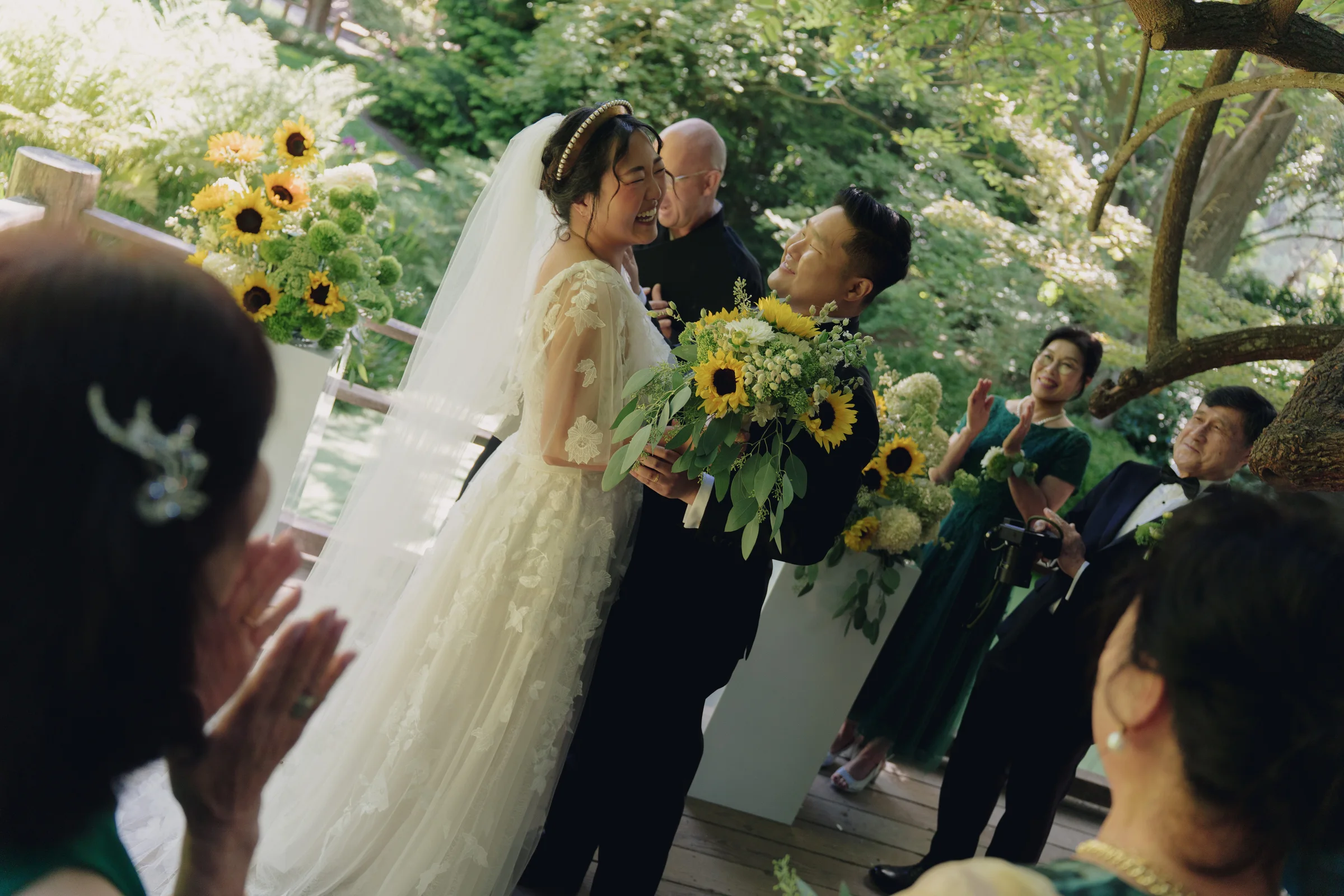 Newlyweds laugh and embrace after their ceremony, surrounded by guests and sunflower arrangements.