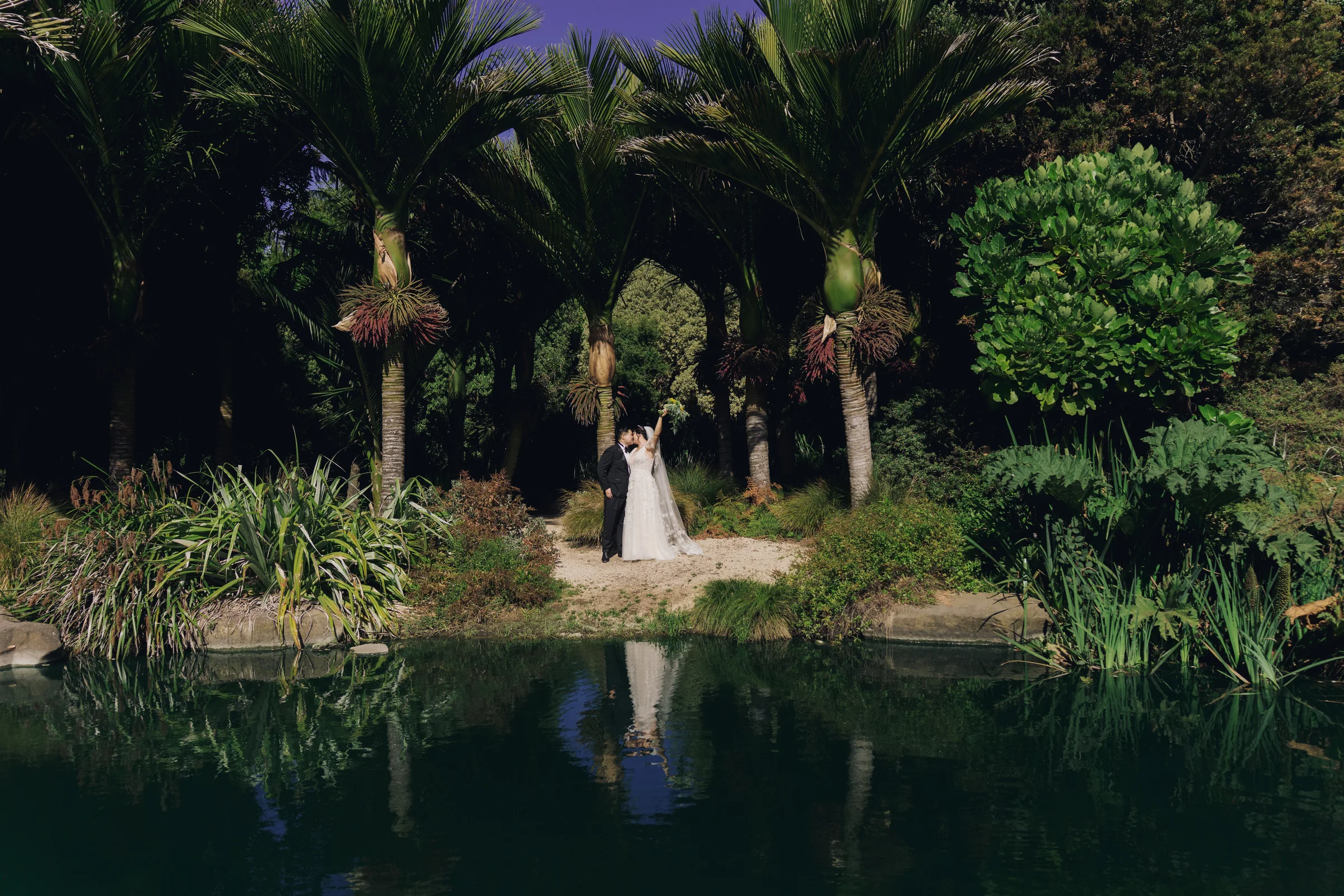 Newlyweds stand among palms beside a reflecting pond as the bride raises her bouquet.