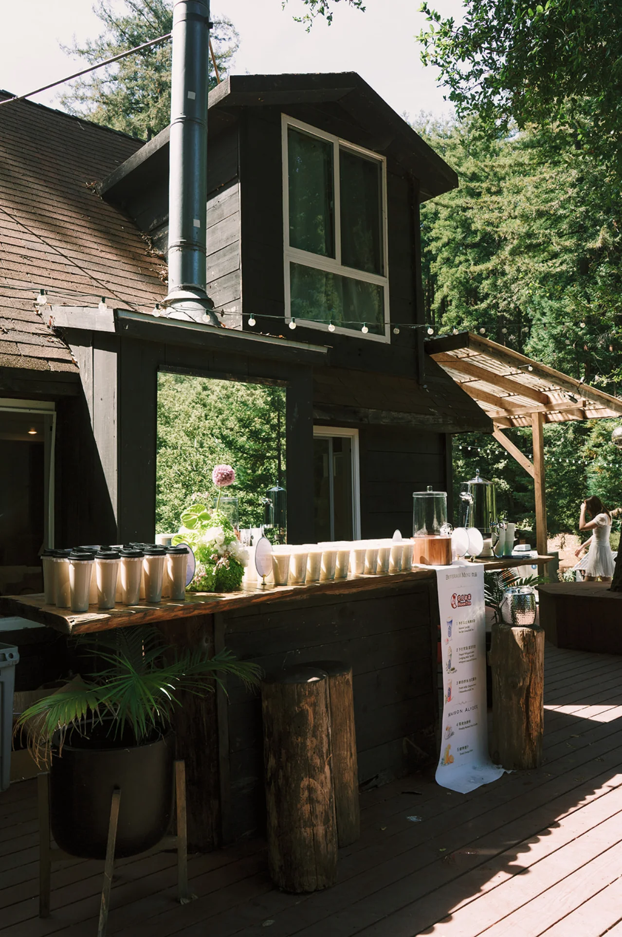 Outdoor milk-tea bar decorated with green and white flowers beside a rustic cabin in the redwoods.