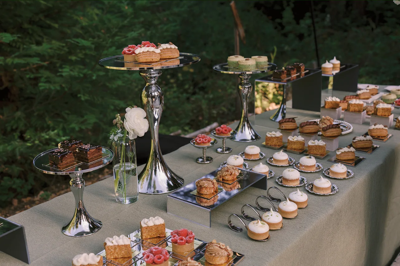 Assorted miniature cakes, tarts, and pastries displayed on mirrored stands with a single white flower.