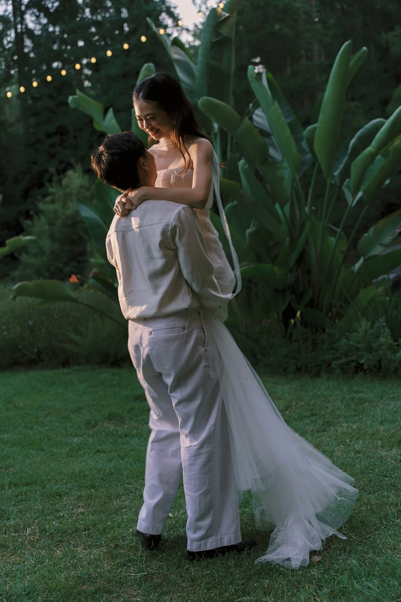 Groom lifting the bride in a garden beneath string lights at dusk.