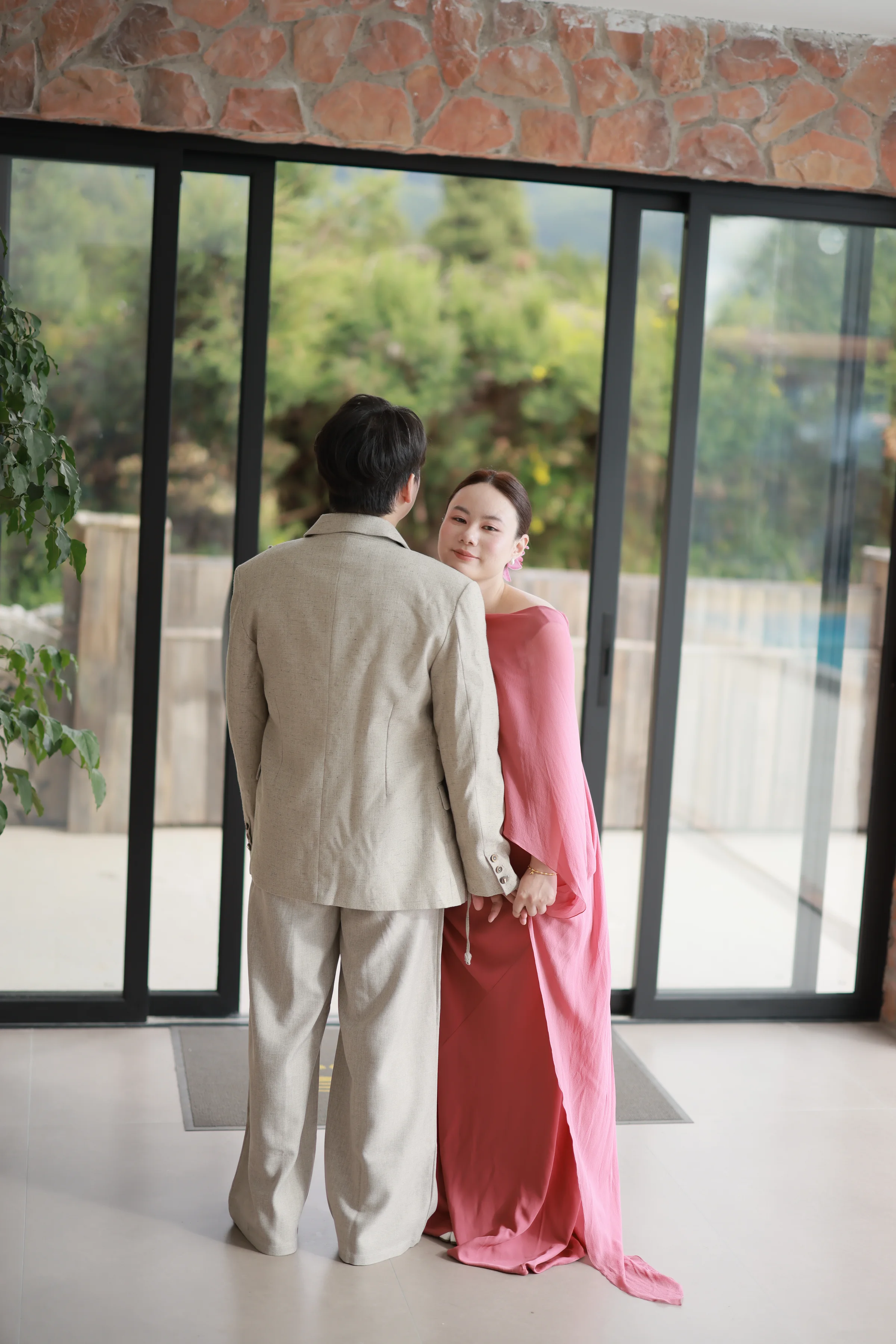 Couple standing by floor-to-ceiling windows, the bride in a flowing coral dress