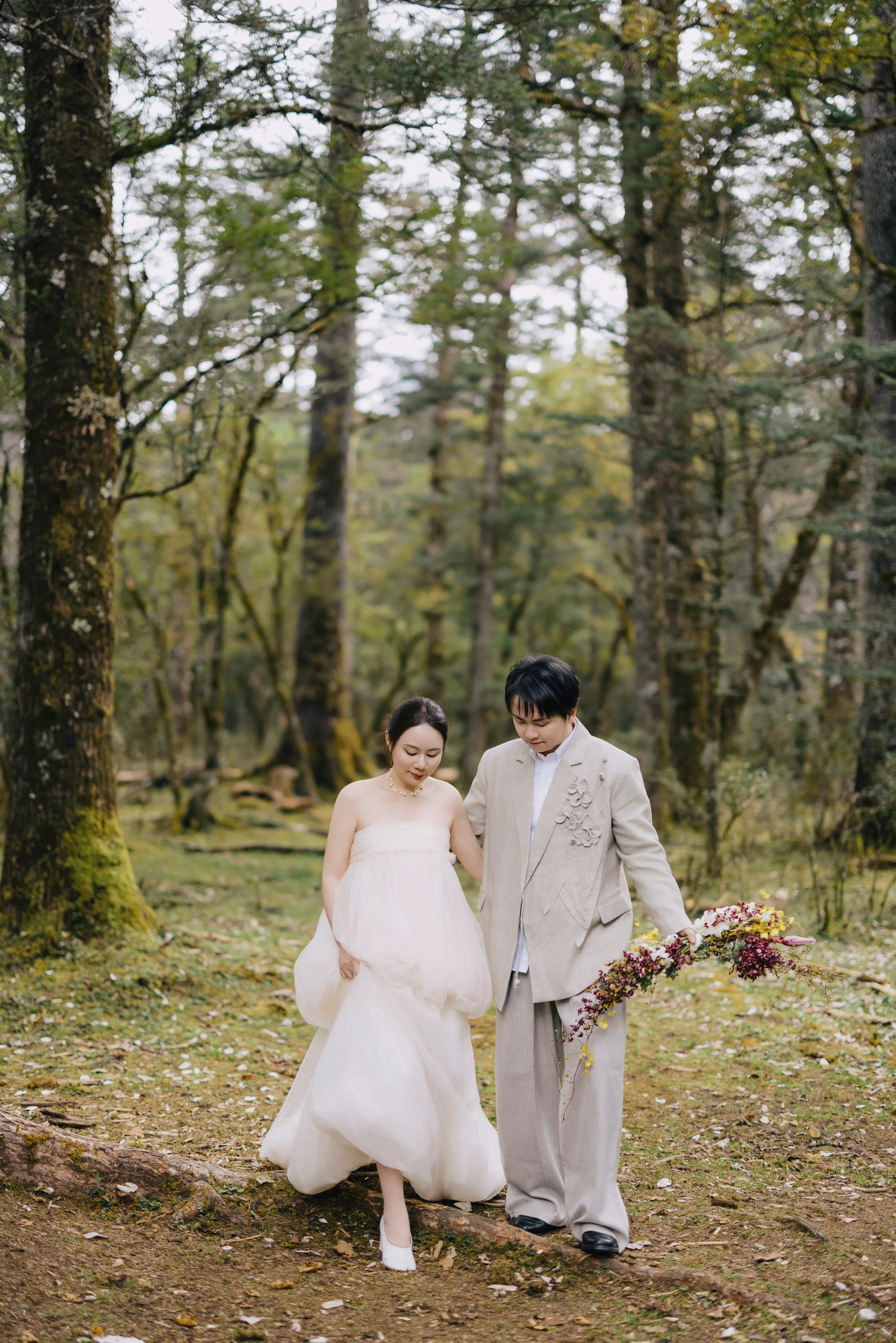 Couple walking hand in hand through a tall pine forest, the bride carrying a bouquet