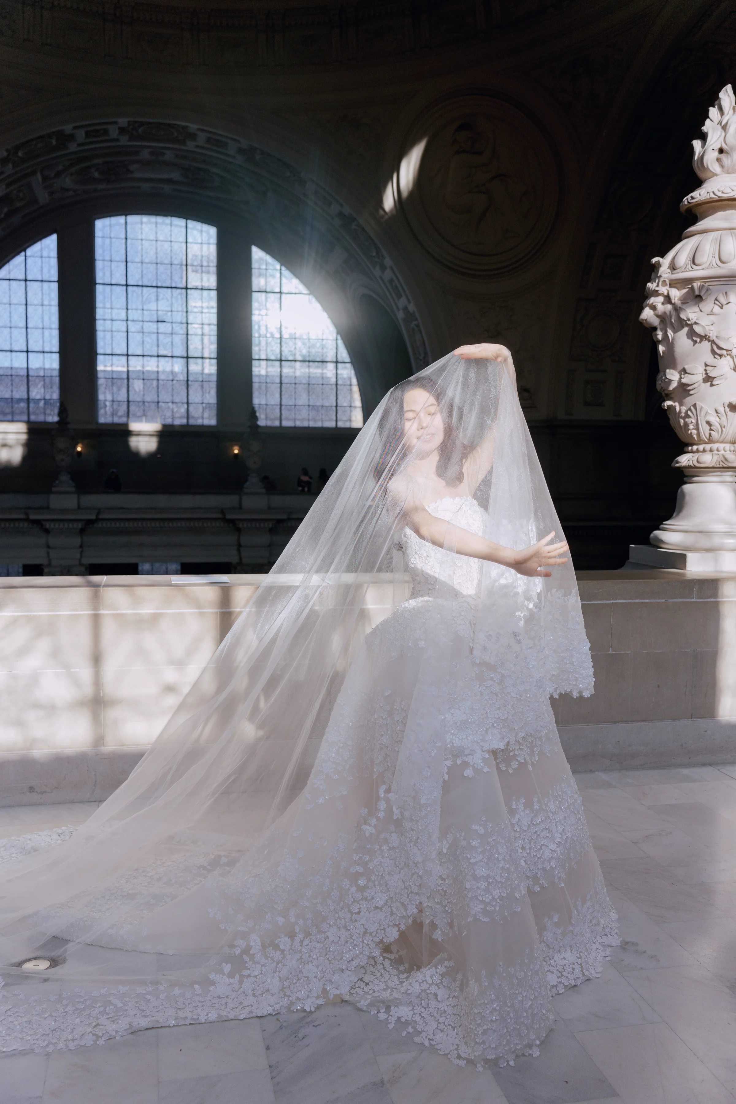 Bride seen from behind, veil and train spread, silhouetted by an arched window