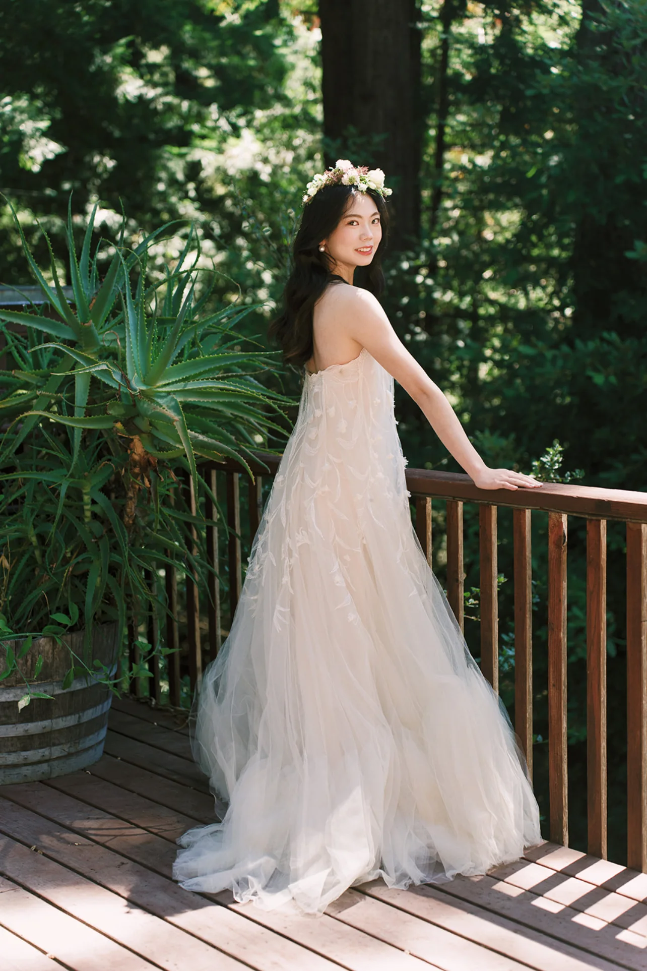 Bride in a flowing floral wedding gown and flower crown standing on a sunlit wooden deck among redwoods.