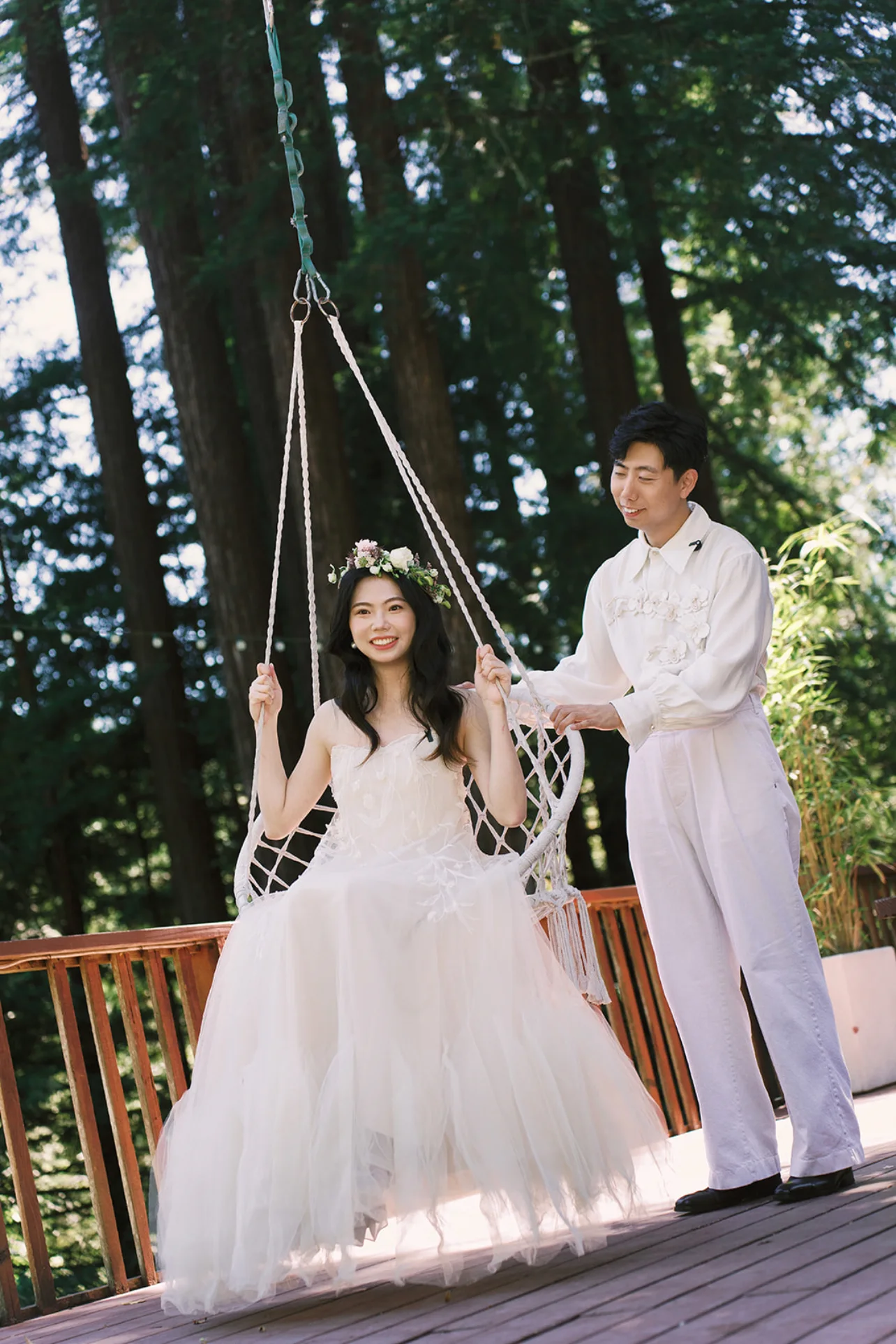 Groom gently pushing the bride on a hanging chair on a deck surrounded by tall redwood trees.