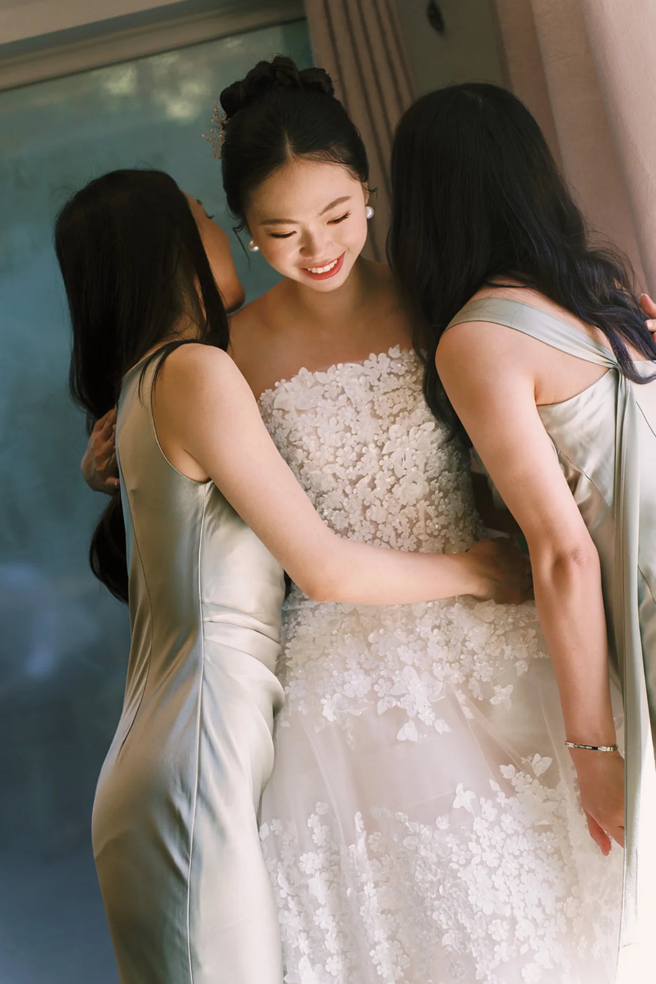 Bride smiling as two bridesmaids in sage green dresses help fasten her floral wedding gown.