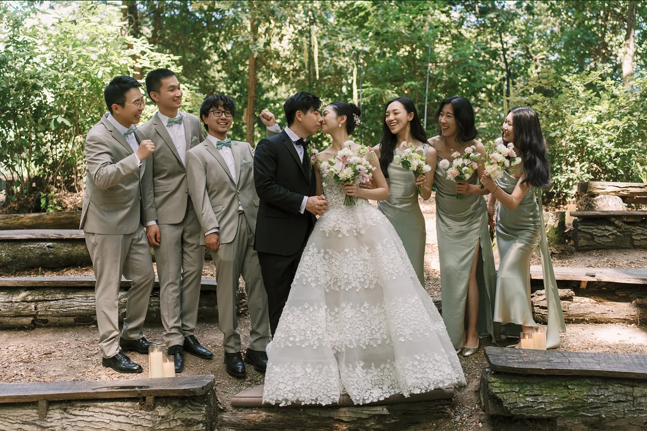 Newlyweds kissing with their wedding party gathered around them in a sunlit redwood grove.