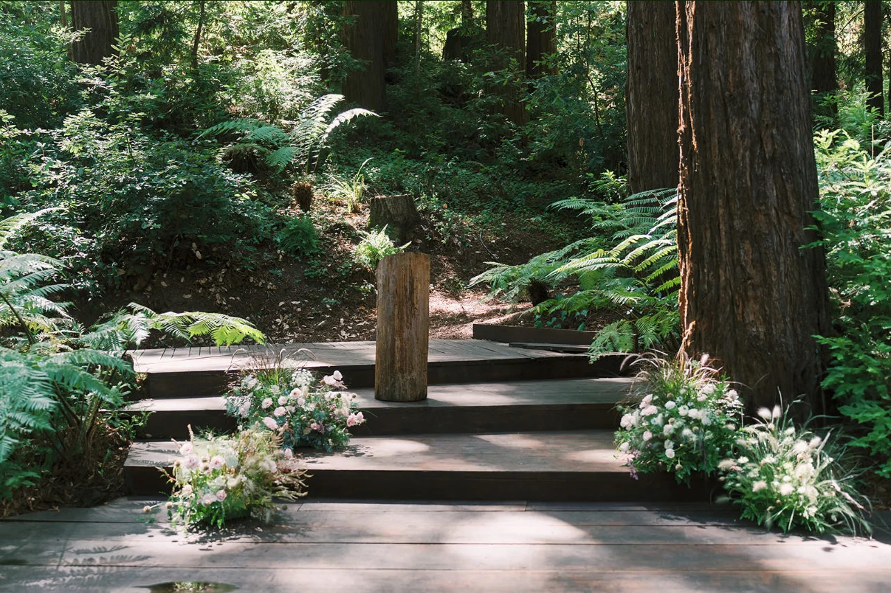 Woodland ceremony platform framed by redwoods, ferns, and low arrangements of pink and white flowers.