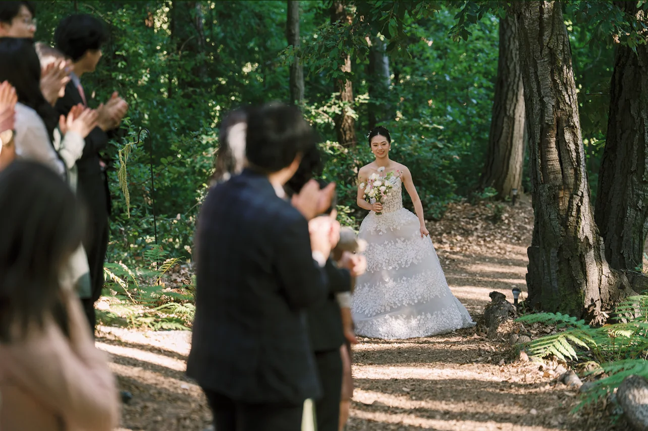 Bride carrying a pastel bouquet down a forest path as guests applaud beneath the redwoods.
