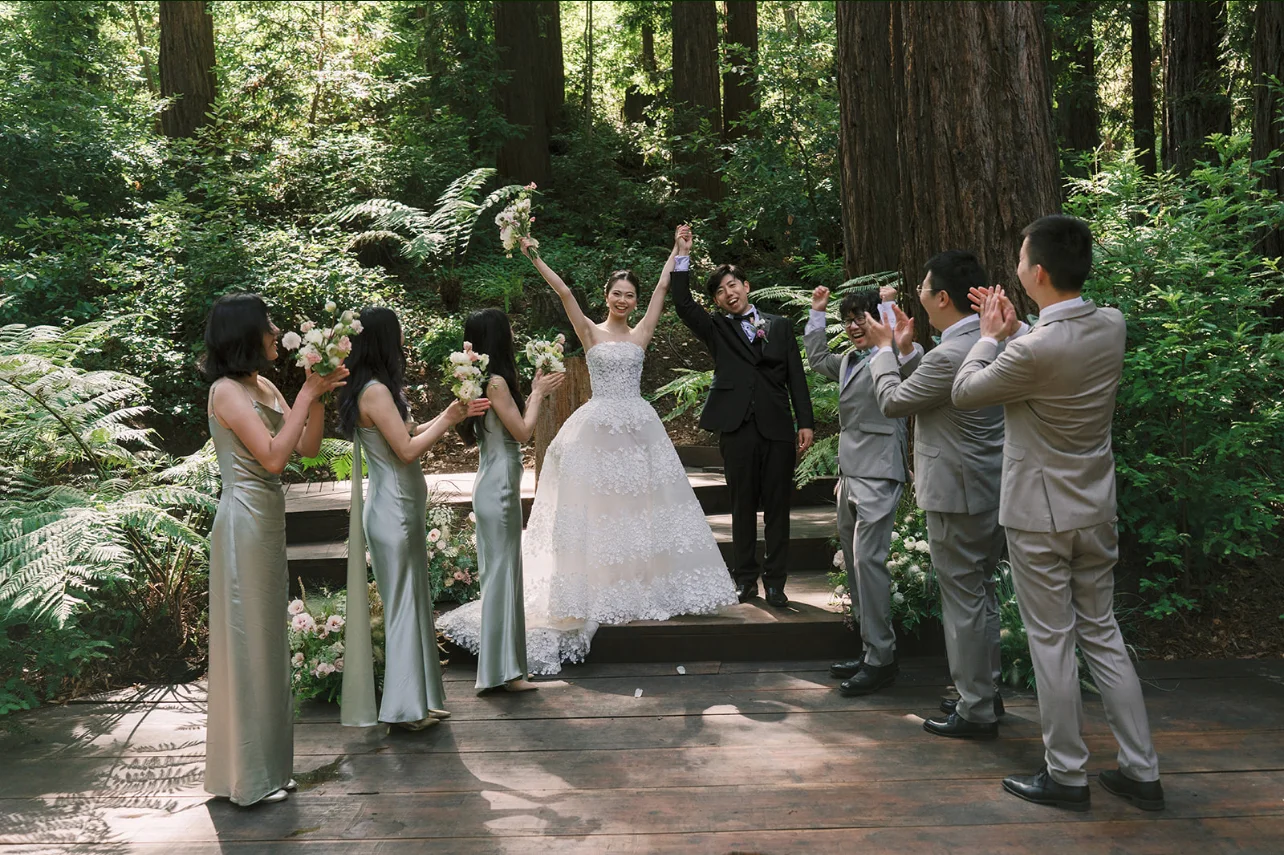 Newlyweds raising their joined hands while their wedding party cheers on the forest ceremony platform.