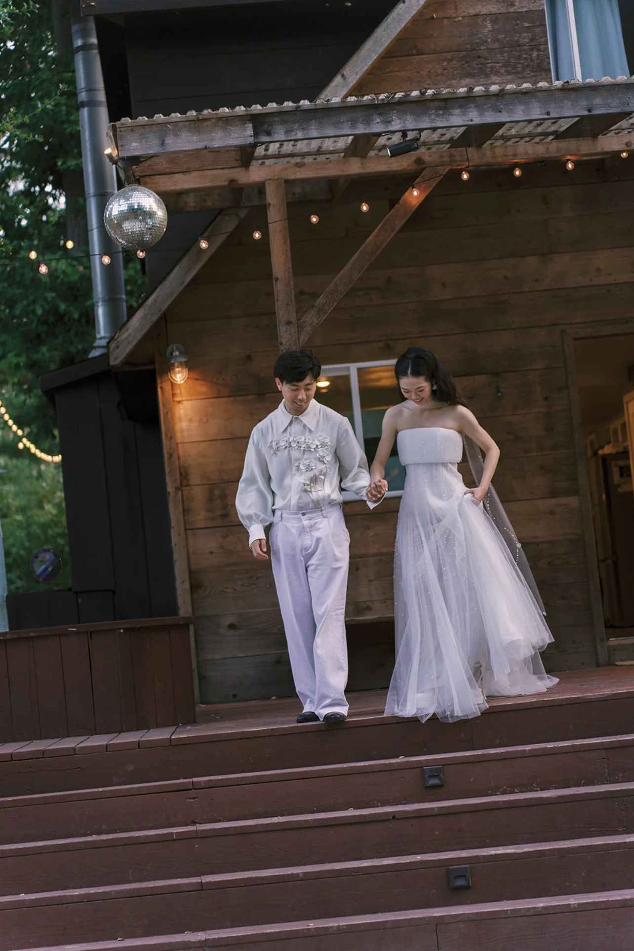 Newlyweds in white reception attire walking hand in hand down the steps of the rustic cabin.