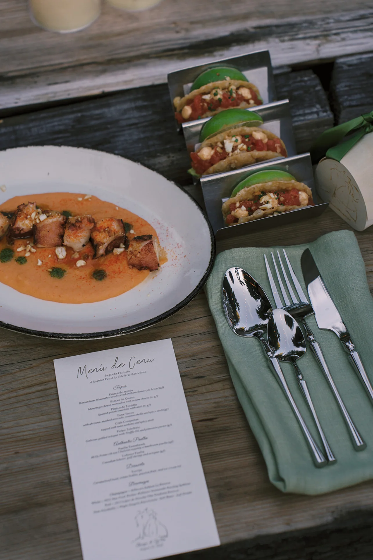 Spanish-inspired dinner plates, small tacos, polished flatware, and a printed menu arranged on a wooden table.