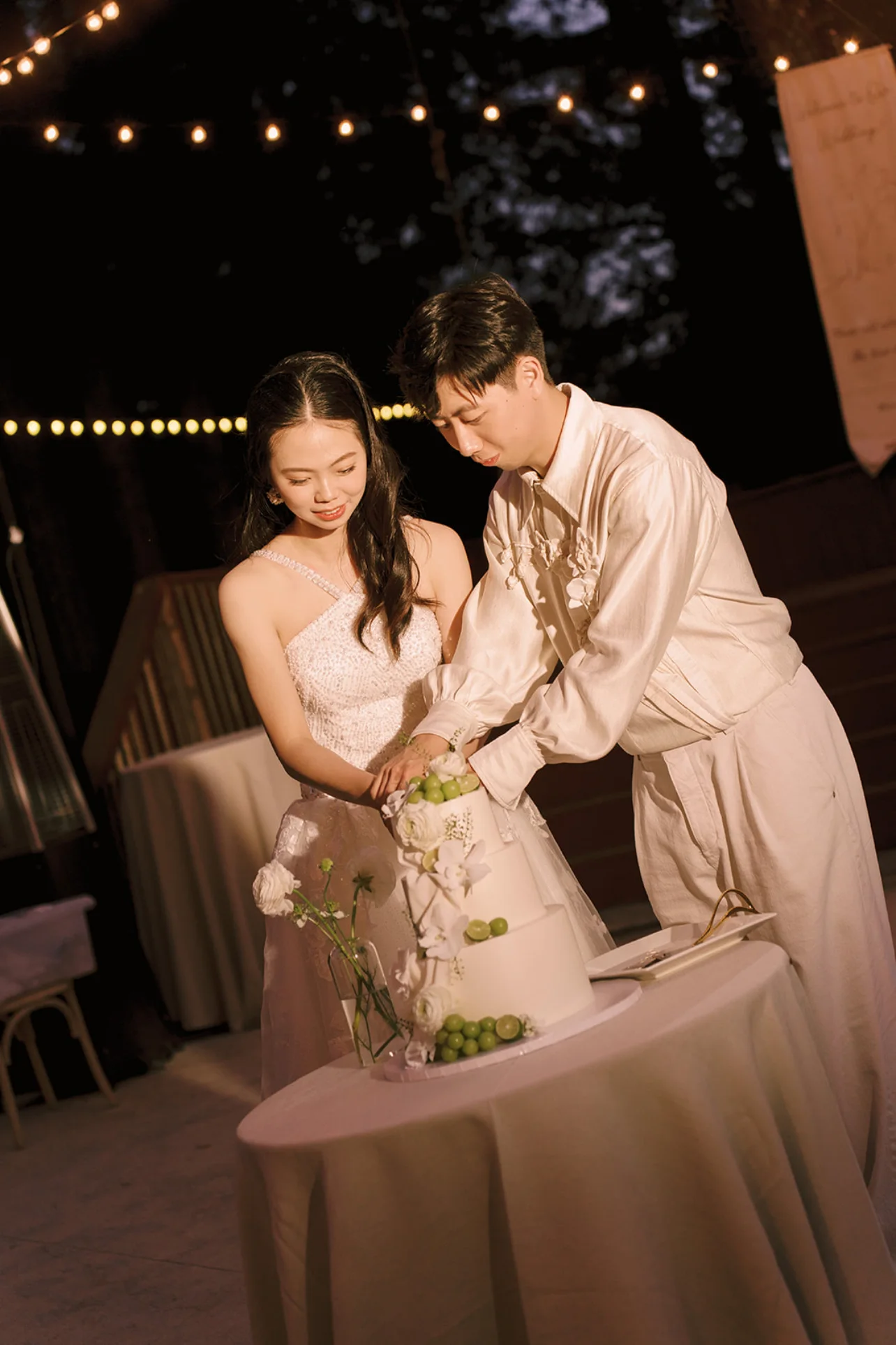 Newlyweds cutting a white wedding cake decorated with green grapes, white flowers, and clear floral tiers.