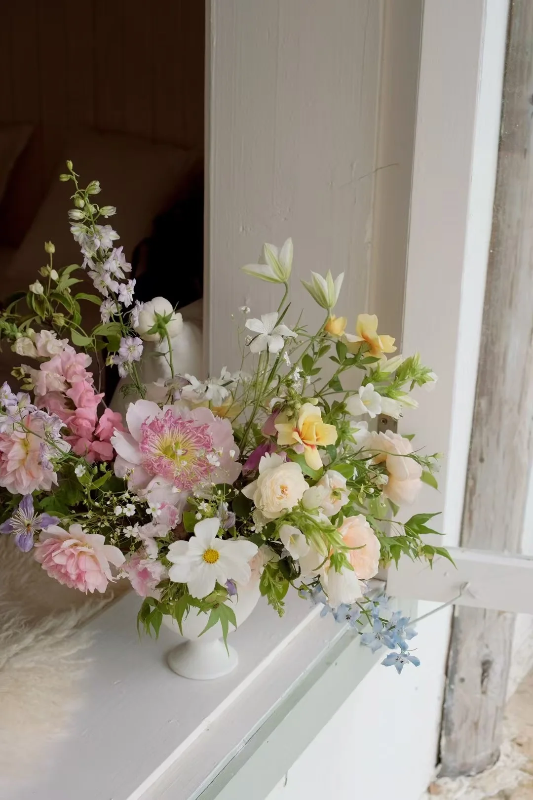 Pastel garden arrangement of peonies, roses, delphinium, and cosmos in a white footed vase on a sunlit windowsill
