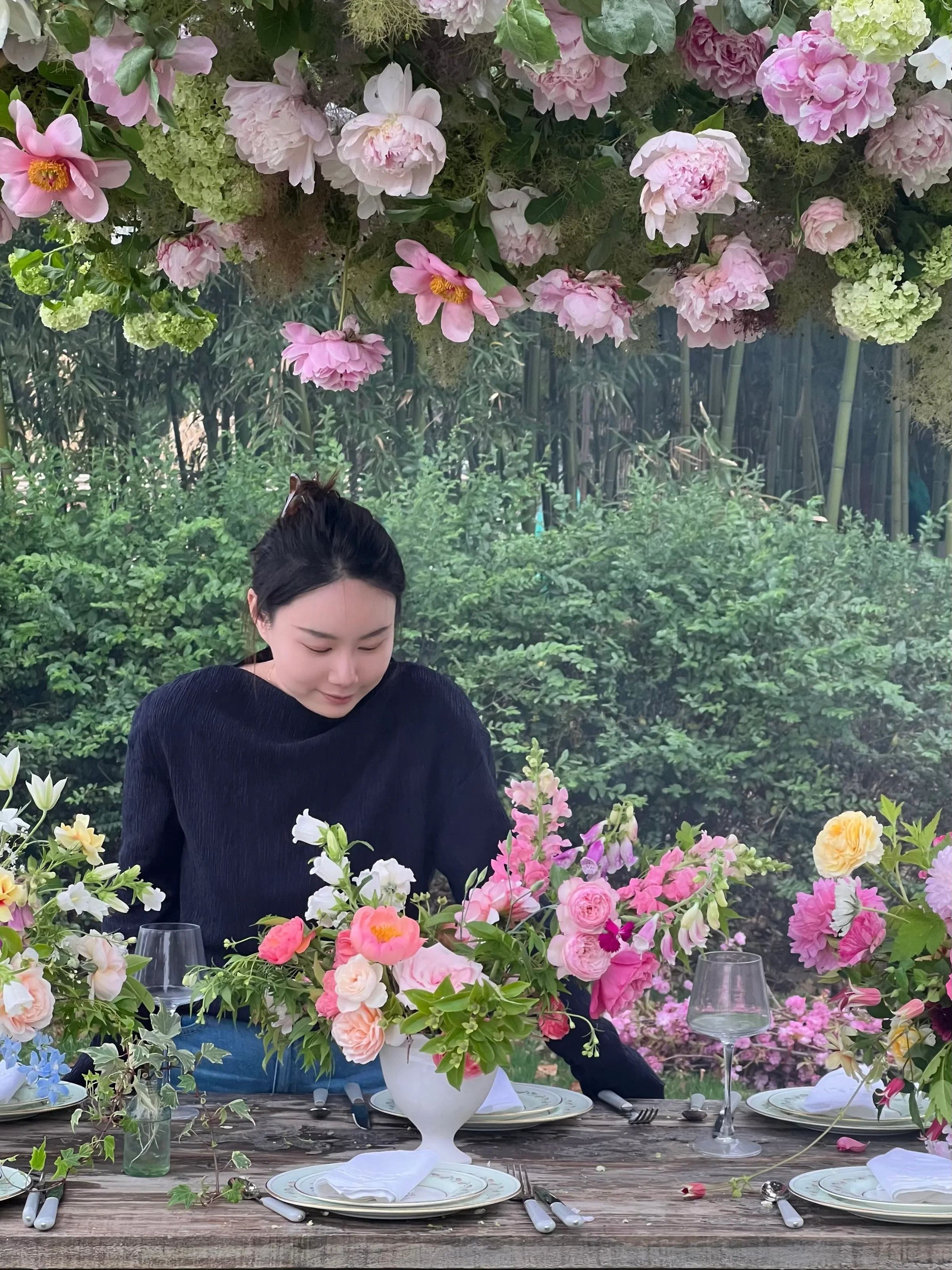 A woman seated at a garden reception table styled with peonies and garden roses beneath a blush peony arch