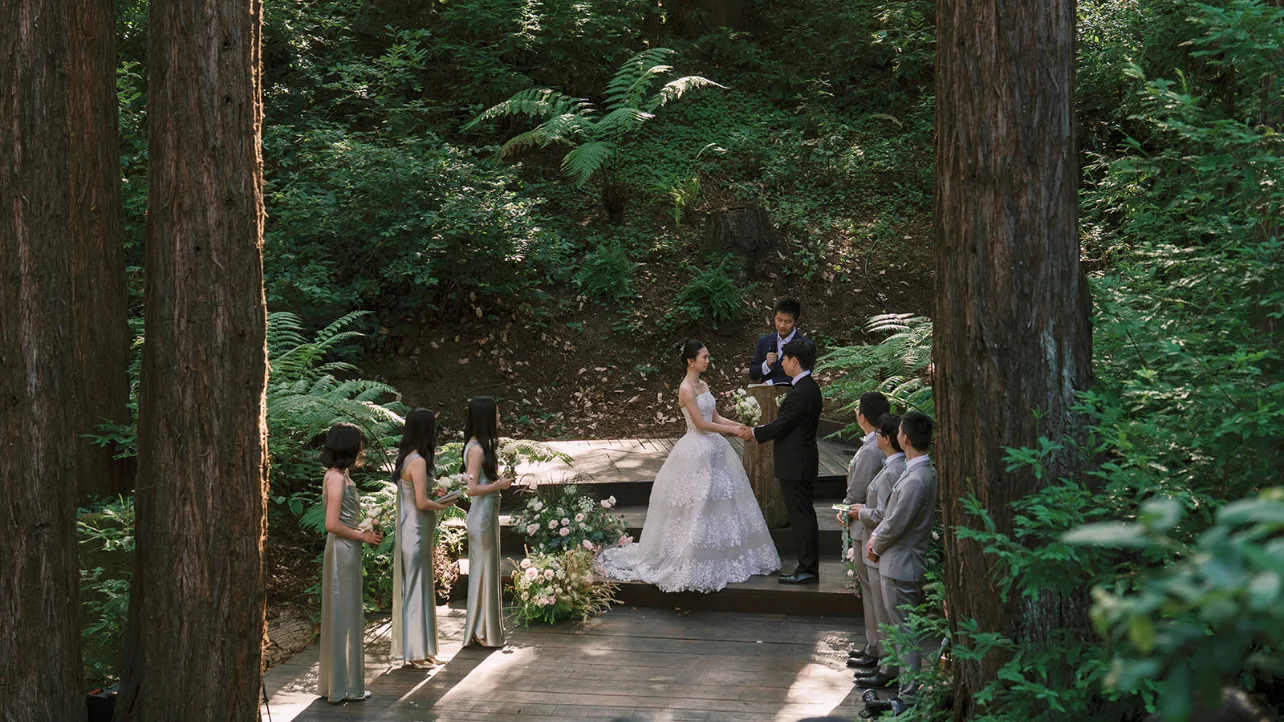 Couple exchanging vows on a woodland deck surrounded by their wedding party and redwoods